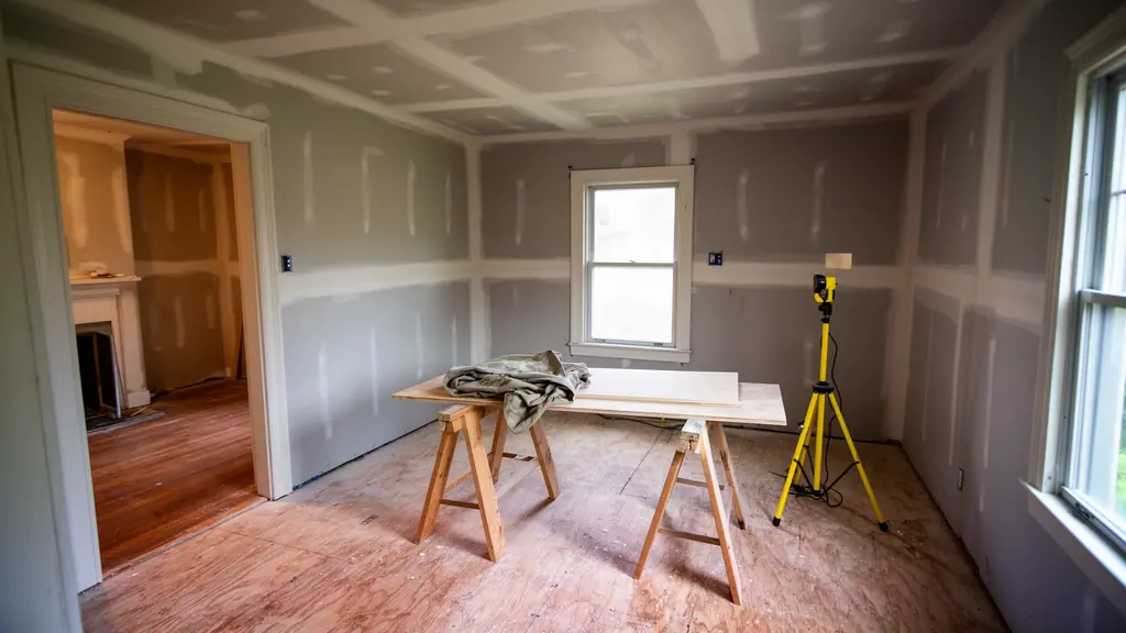 Room mid-remodel with new unpainted drywall, a plywood subfloor, sawhorses holding a sheet of plywood and a tripod work light.